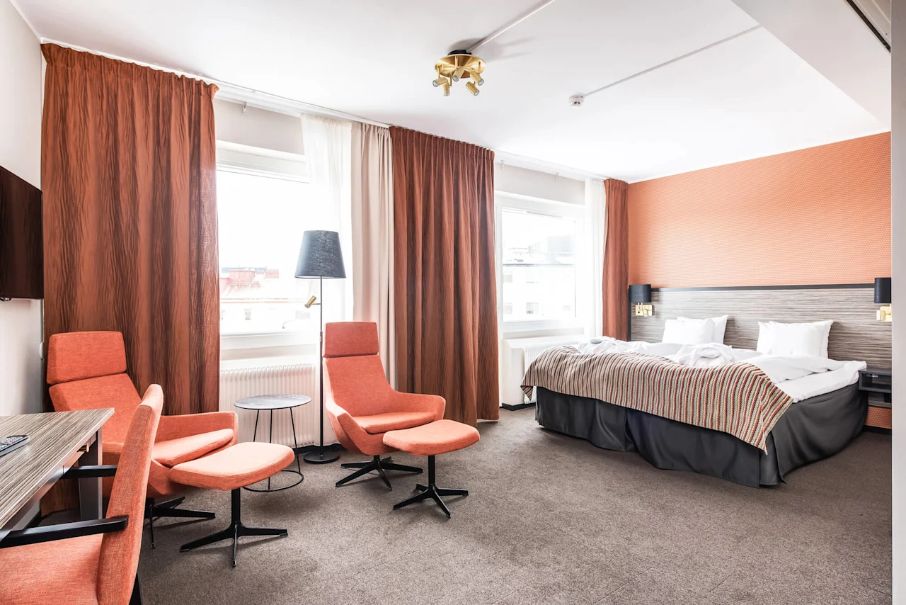 Et velopplyst hotellrom med seng, sitteområde og store vinduer.
