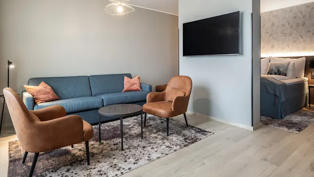 En blå sofa med oransje puter står overfor to brune lenestoler og et rundt bord på et mønstret teppe. En TV er montert på veggen; et soverom er delvis synlig.