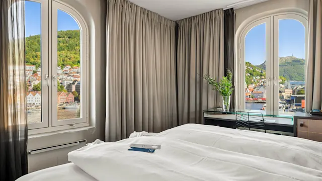 A neatly made bed with white linens is beside two arched windows showing a picturesque town and green hills. A desk with a plant sits nearby.