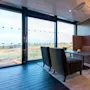 A cozy seating area with a long couch, two armchairs, a table, and a lamp. Large windows provide a scenic view of a grassy landscape outside.