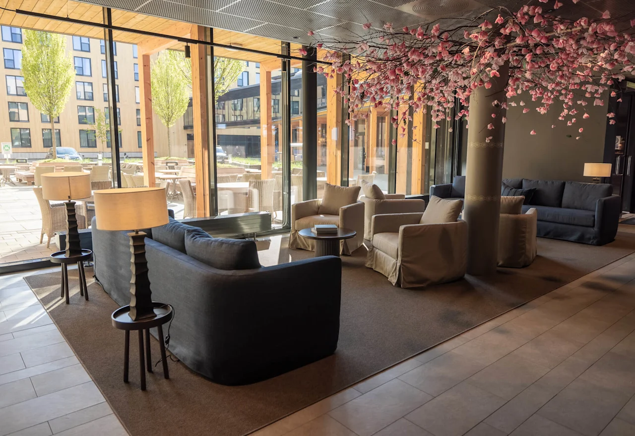 En moderne loungeområde med sofaer, stoler og et dekorativt kirsebærtre over. Store vinduer viser en uteveranda og urban bakgrunn.
