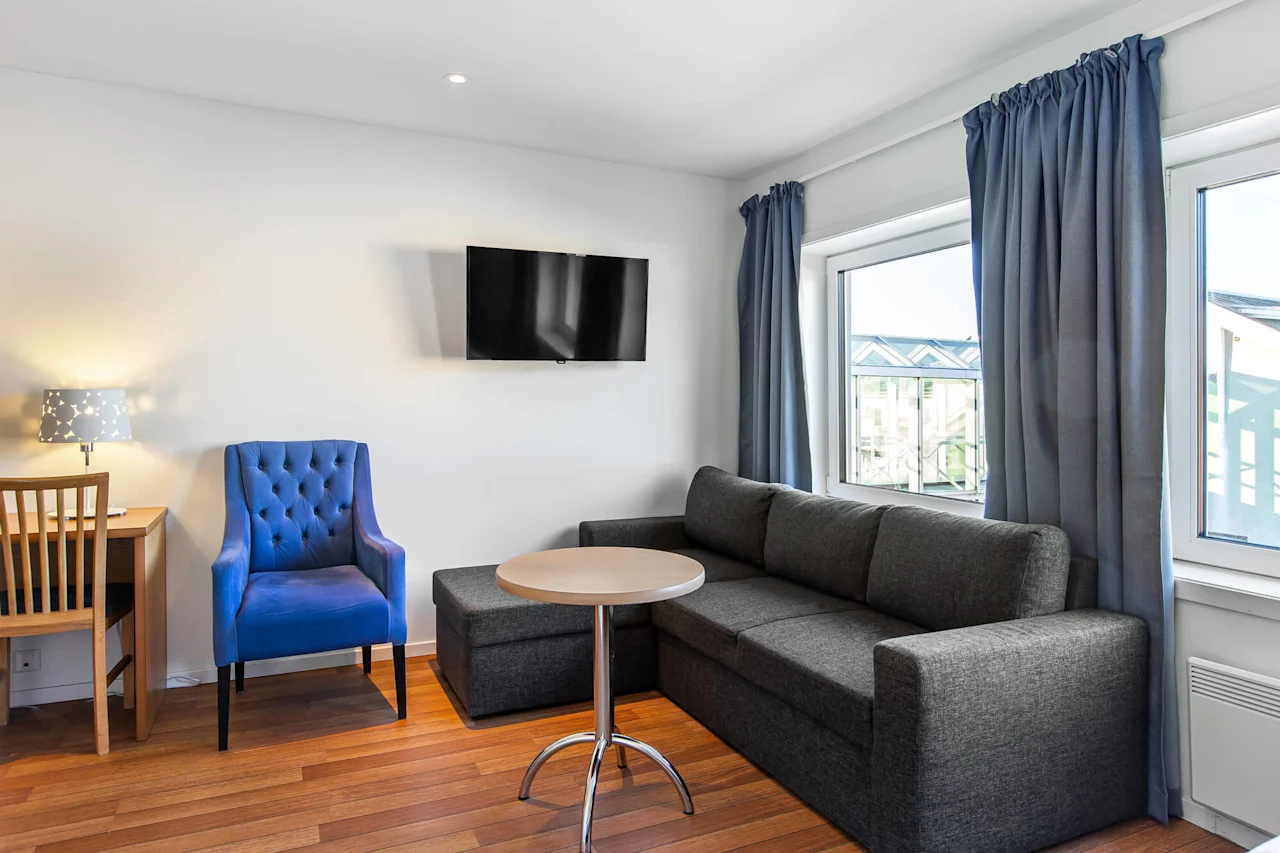 A modern living room features a dark gray sofa with a round table, a blue armchair, a wall-mounted TV, and a wooden desk with a lamp, framed by large windows and curtains.