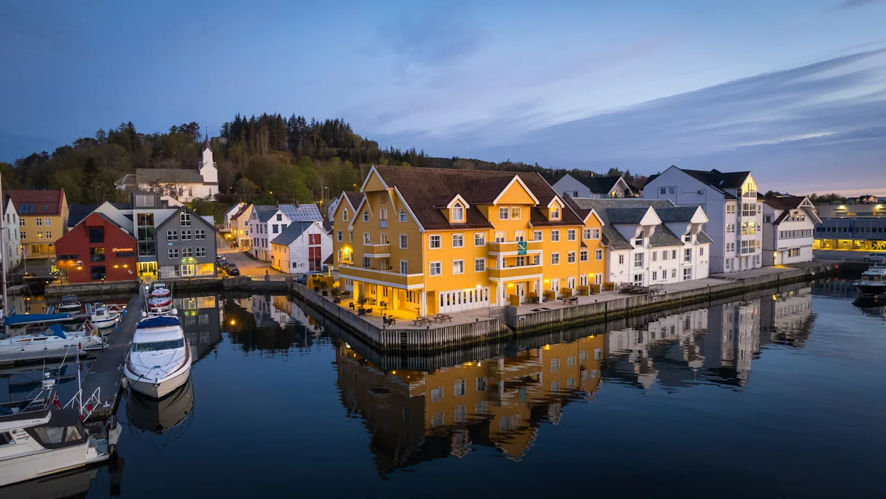 Gul bygning ved vandet afspejler sig i roligt aftenhav, omgivet af andre farverige bygninger og både. En kirke og træer er synlige i baggrunden under en aftenhimmel.