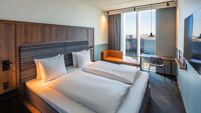 Et hotellrom inneholder to pent reddede senger. Ved siden av dem står et skrivebord og stol ved et stort vindu med utsikt over byen. Veggmontert TV er til stede.