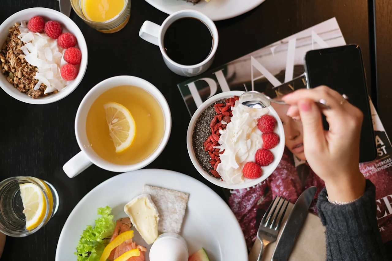 En hånd holder en skje over en frokostbolle med chiafrø, kokos, gojibær og bringebær ved siden av te, juice og et magasin på et mørkt bord.
