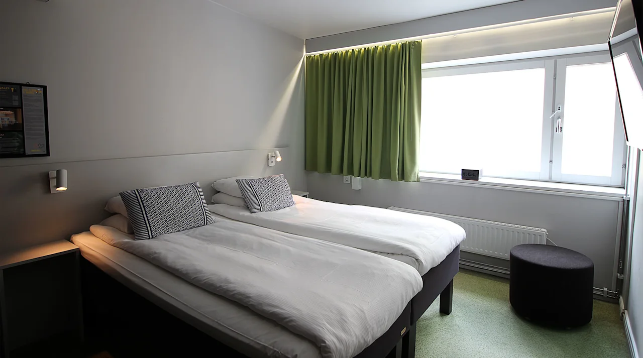 To pent oppredde enkeltsenger med mønstrede puter er i et minimalistisk rom. Et stort vindu med grønne gardiner gir naturlig lys. En liten rund puff er nær radiatoren.