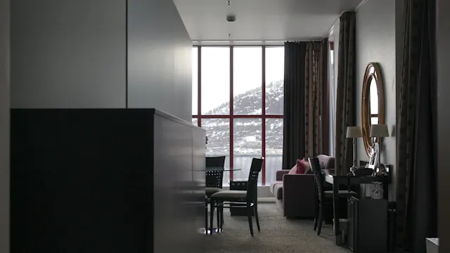 Et moderne og svakt opplyst hotellrom med møbler, utsikt over snøkledde åser og vann gjennom store vinduer.