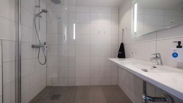 En moderne dusj med glassvegg, ved siden av en bred hvit servant med stor speil i et flislagt baderom. Lys belysning lyser opp det rene, minimalistiske rommet.