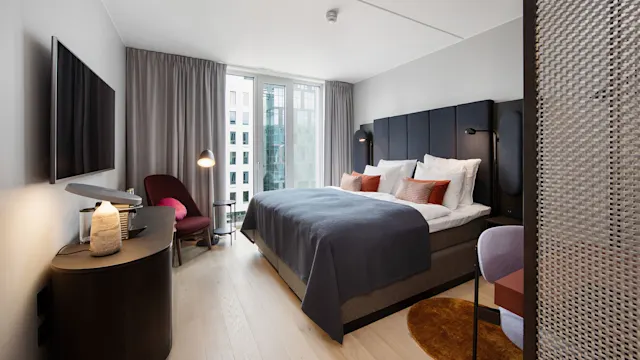 A neatly made bed with blue bedding, adorned with white and orange pillows, sits beside large windows in a modern hotel room, featuring a chair and television opposite.