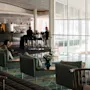 Komfortable loungestoler med puter er plassert nær glassvegger, mens en kvinne drikker kaffe. Bak er en moderne bar med sittende personer synlig i et lyst miljø.