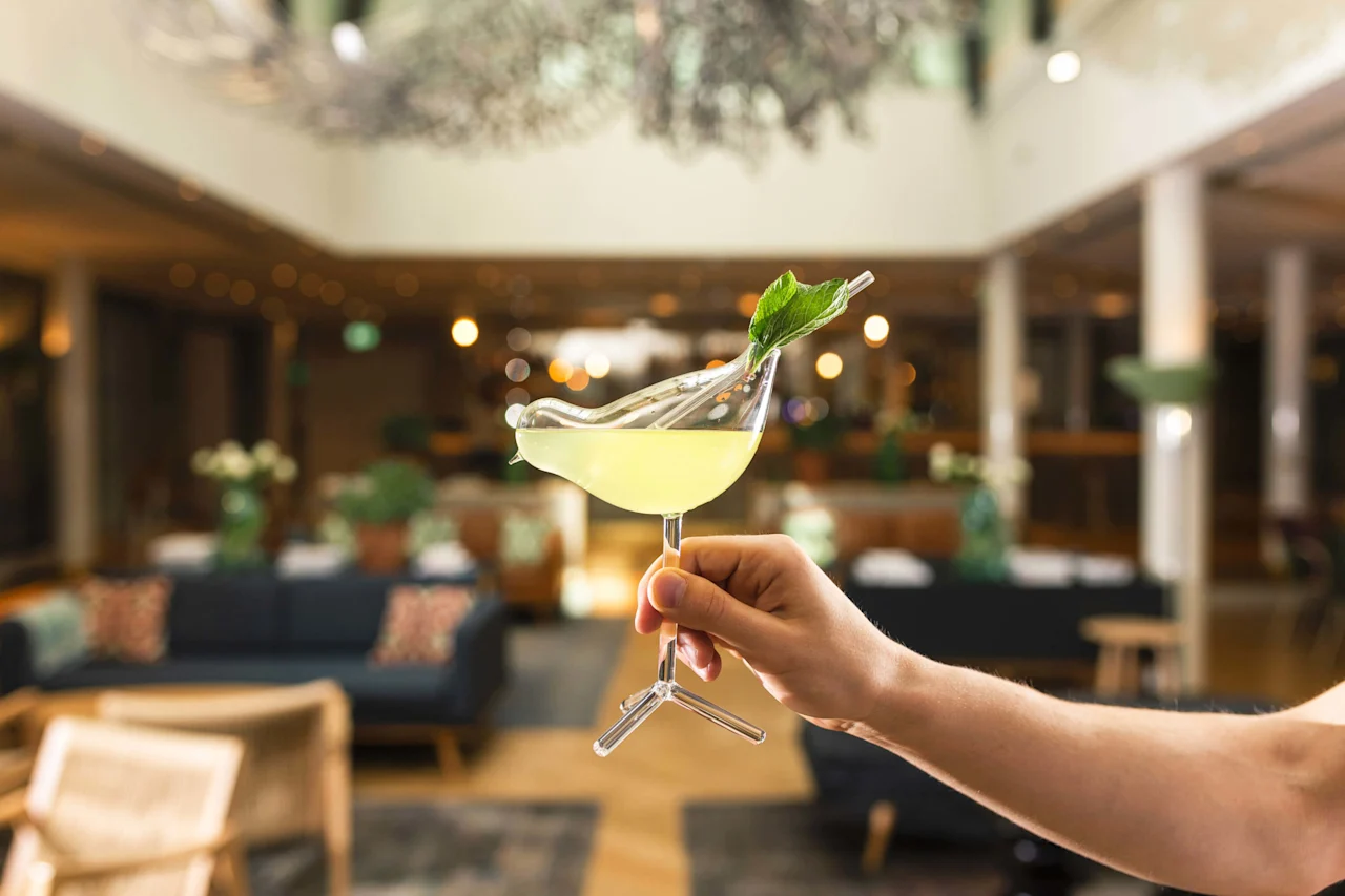 Käsi esittelee cocktailin mintunlehden koristeella, taustalla sumea ylellinen baarimiljöö.