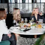 Fyra kvinnor sitter och pratar vid ett runt bord, njuter av drycker och olika rätter i en elegant restaurangmiljö. Lummiga växter och modern inredning omger dem.