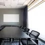 Et langt svart konferansebord omgitt av svarte stoler står i et møterom med en whiteboard på en grå vegg. Store vinduer med gardiner gir naturlig lys.