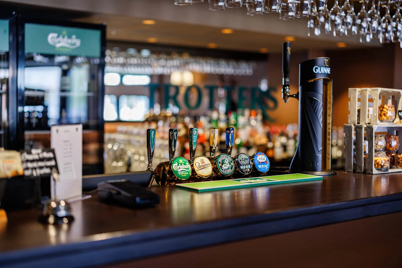 Ölkranar med olika märkesetiketter är uppradade på en bardisk i trä i en pub. Ovan hänger glas upp och ner. I bakgrunden syns en suddig bardisk. Text: "Guinness."