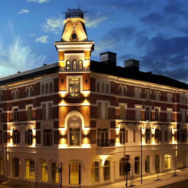 A multi-story building with arched windows is illuminated at night. It stands on a street corner, surrounded by similar architecture, under a twilight blue sky.