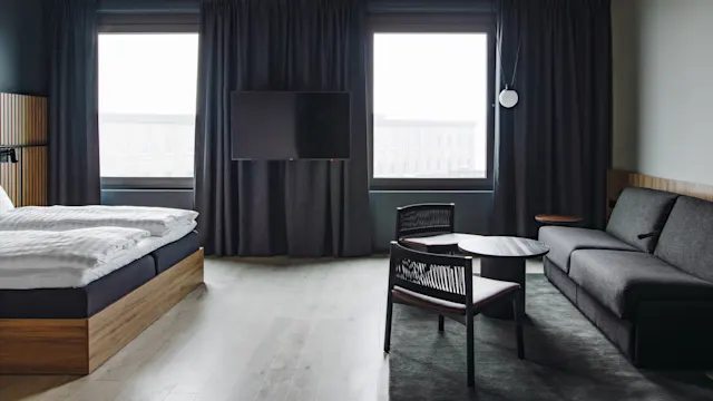 Bed covered with white linens; stands beside a window with dark curtains. A television is mounted above. Nearby, a gray sofa and two chairs surround a small table on the floor.