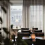 Spiseborde er arrangeret med stole, centreret omkring et lysende lys i en blødt oplyst restaurant. Store gardiner dækker vinduer, delvist åbnede mod bygninger udenfor.