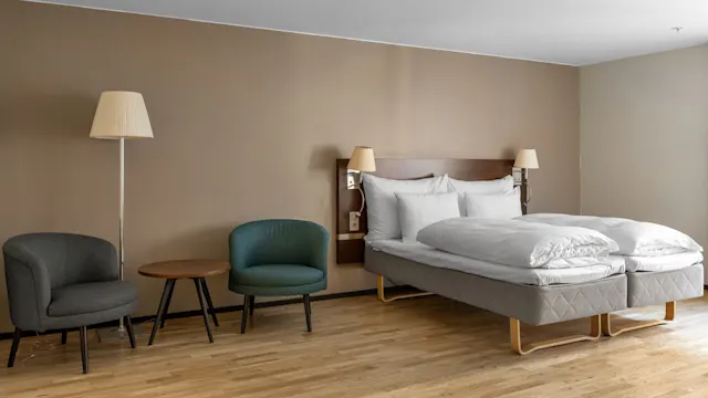 A bed with white linens stands against the wall, flanked by lamps, next to two chairs and a small round table on a wooden floor.