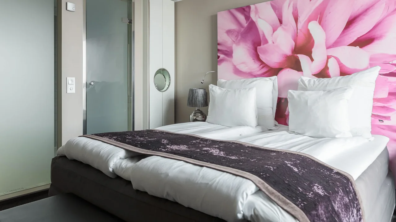 En seng med hvite puter og et mørkt teppe står mot en vegg med et stort rosa blomstermotiv. En lampe og en frostet glassdør er i nærheten.