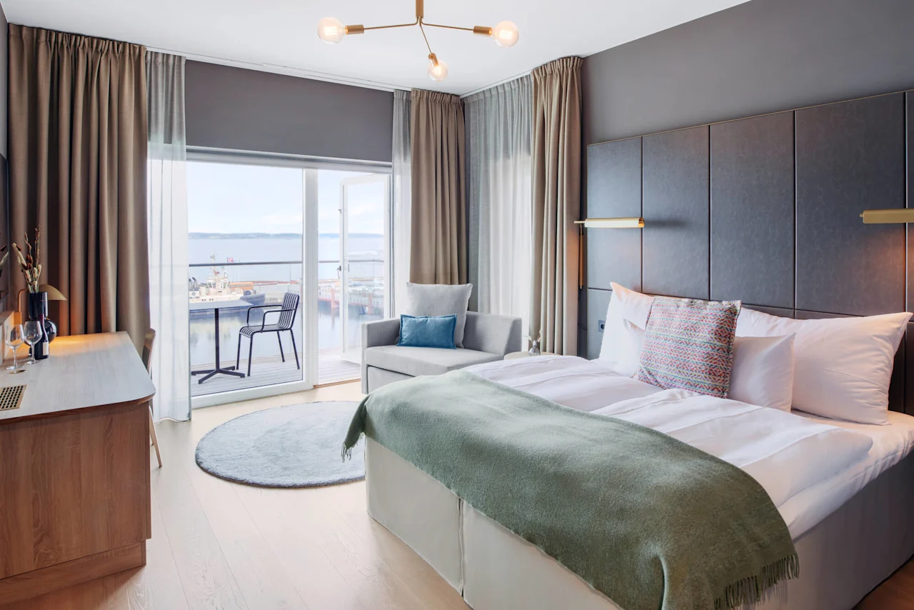 Et pent oppstilt hotellrom har en stor seng med puter og teppe, et skrivebord med glass, og en grå sofa. Balkongdørene åpner mot en vannutsikt med stoler utenfor.