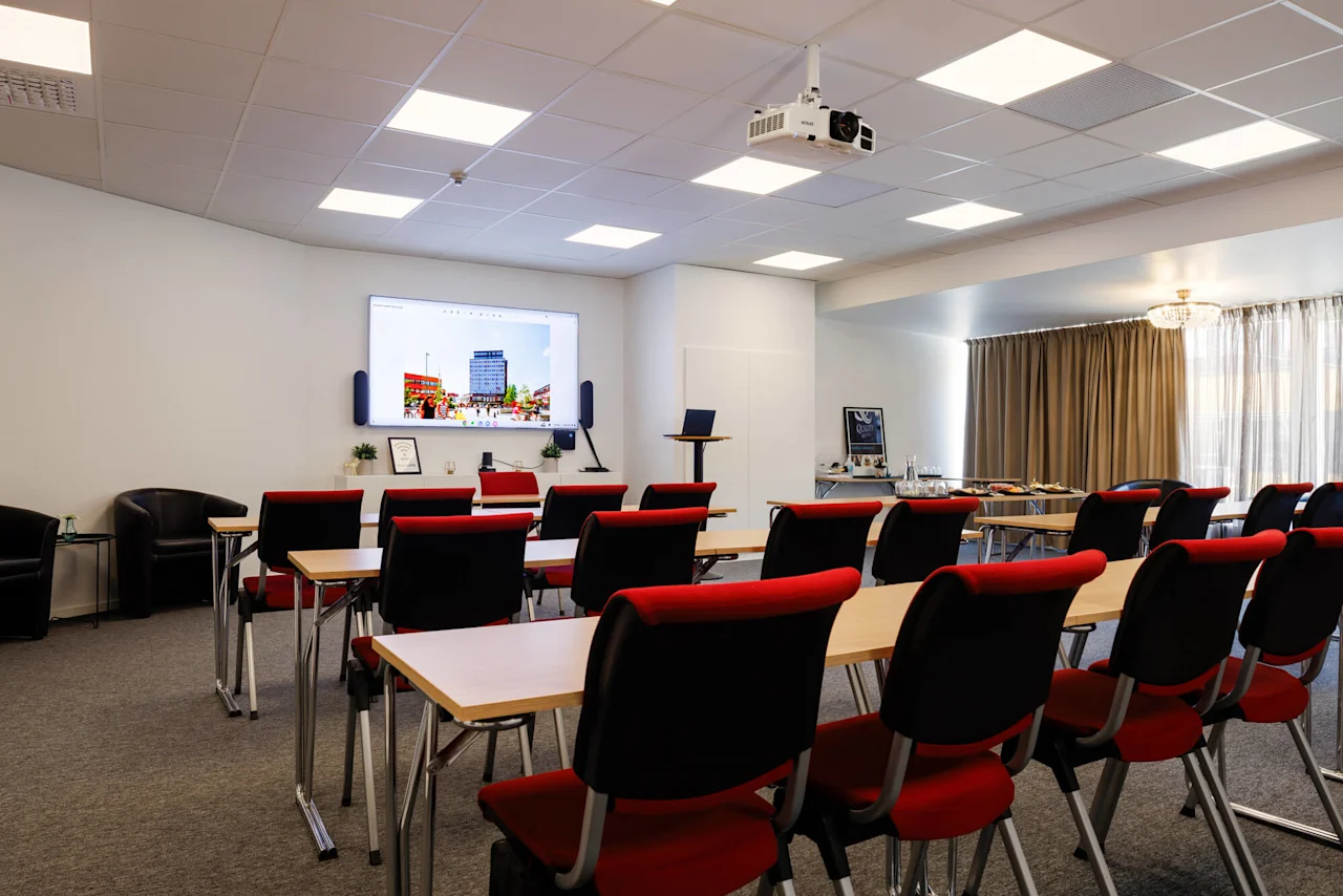 Konferensrum med rader av stolar vända mot en skärm som visar en stadsbild. En projektor är monterad i taket, och bord med förfriskningar finns bak.