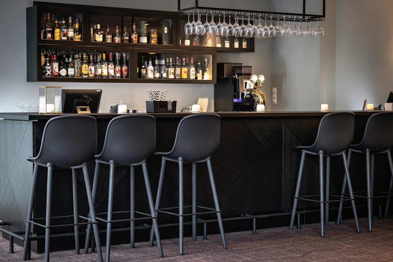 En elegant bar med høye stoler står foran hyller med pent organiserte spritflasker. Glass henger over, og myk belysning skaper en koselig atmosfære.