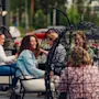 En gruppe kvinner sitter og prater i hengestoler utendørs. De smiler, omgitt av grøntområder og parkerte biler, som antyder en avslappet sosial sammenkomst i hageaktige omgivelser.