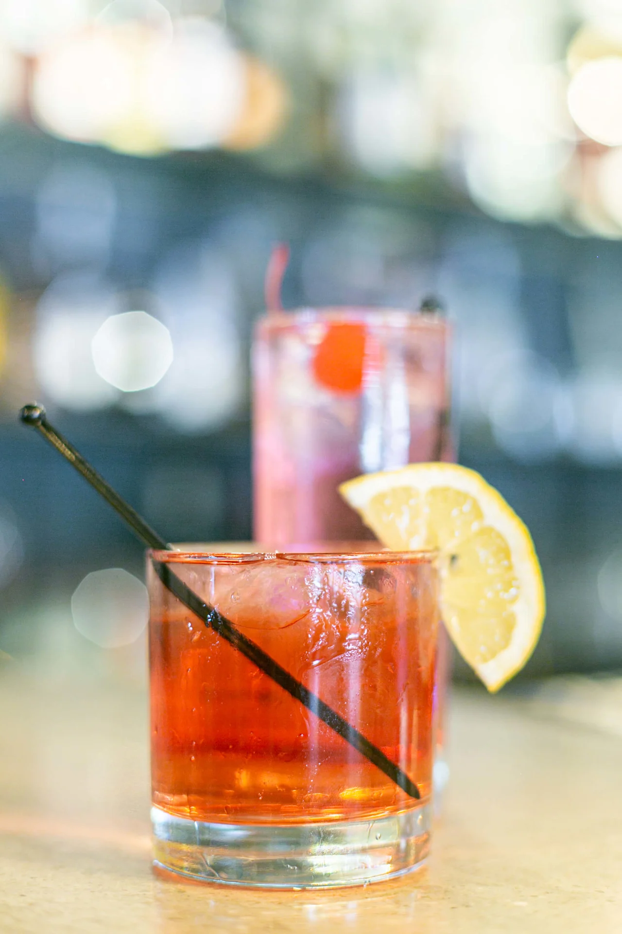 En röd cocktail med is och en citronskiva på kanten står i ett lågt glas med ett svart sugrör, mot en suddig barbakgrund.