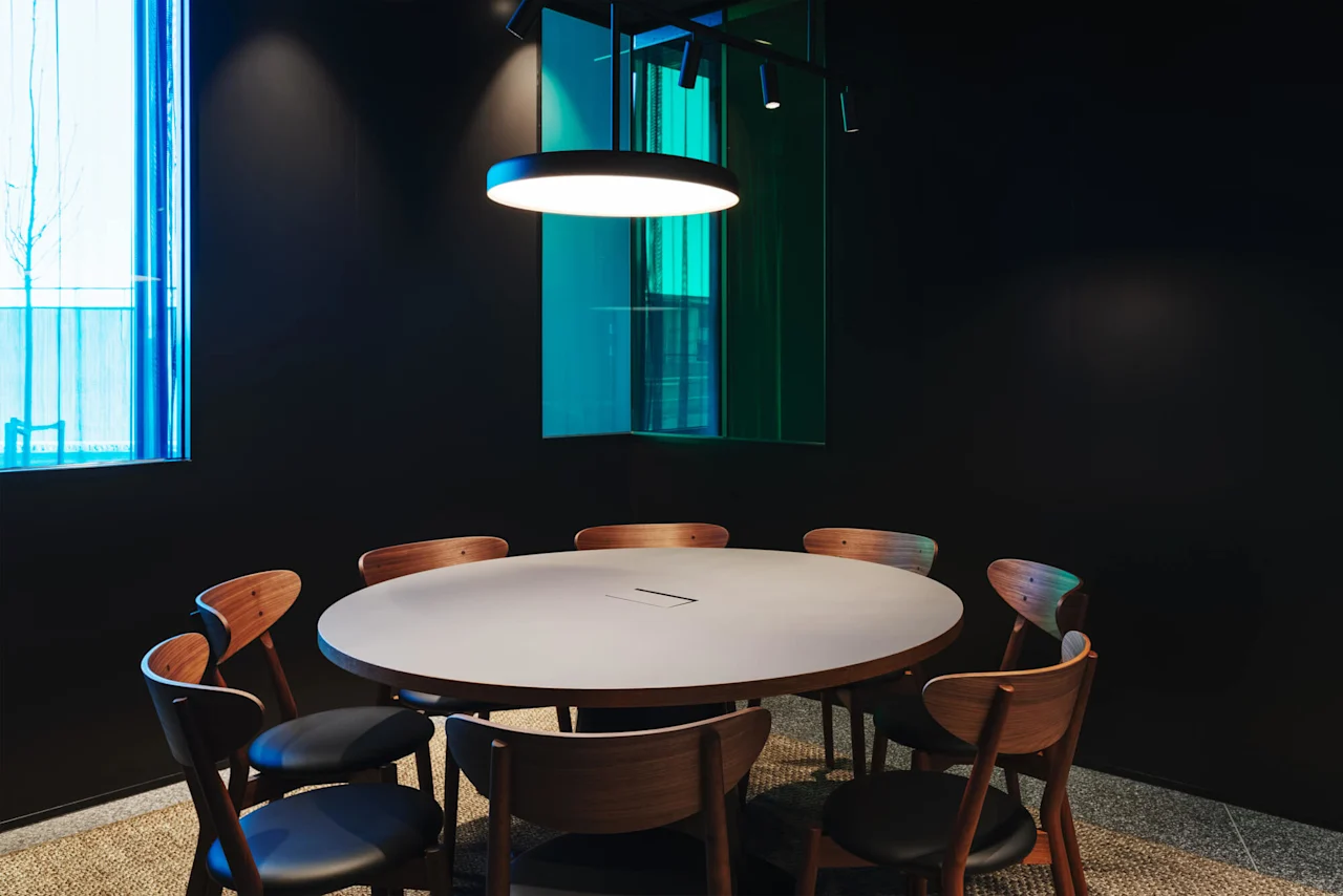 A round table surrounded by wooden chairs sits under a hanging light in a dimly lit room with a blue-tinted window.