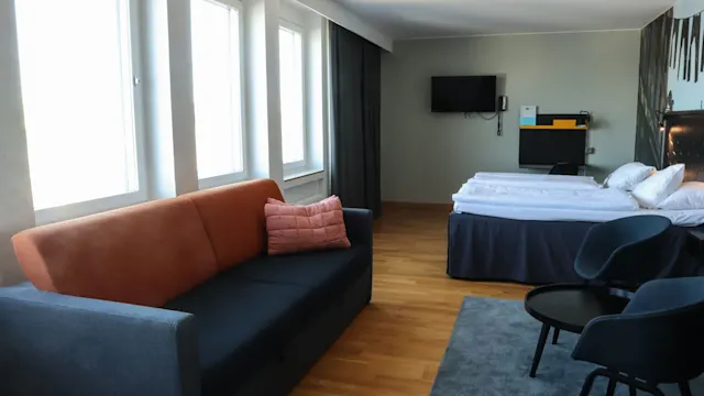 En mjuk orange soffa med en rosa kudde står i ett modernt hotellrum. I närheten finns en säng och en stol på trägolv under ljusa fönster.
