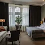 En seng med hvitt sengetøy og et grått teppe flankeres av lamper på trebenker; en lenestol og sofa står i nærheten, med et stort vindu og gardiner i bakgrunnen.