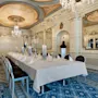 A long, elegantly set dining table is situated under ornate chandeliers in a luxurious, mirrored room with intricate ceiling designs and blue patterned carpet. No text is present.