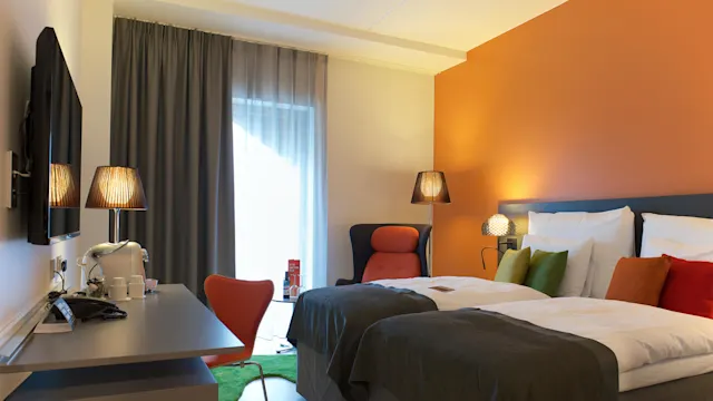 Ett modernt hotellrum har två enkelsängar med färgglada kuddar, sänglampor och ett skrivbord med en stol. Ett fönster med gardiner ger naturligt ljus. En vägg är målad i orange.