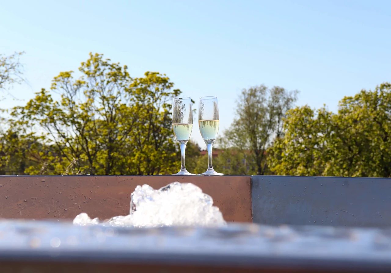 To champagneglass, plassert på en våt overflate med boblende skum, omgitt av et utendørsmiljø med grønne trær i bakgrunnen.