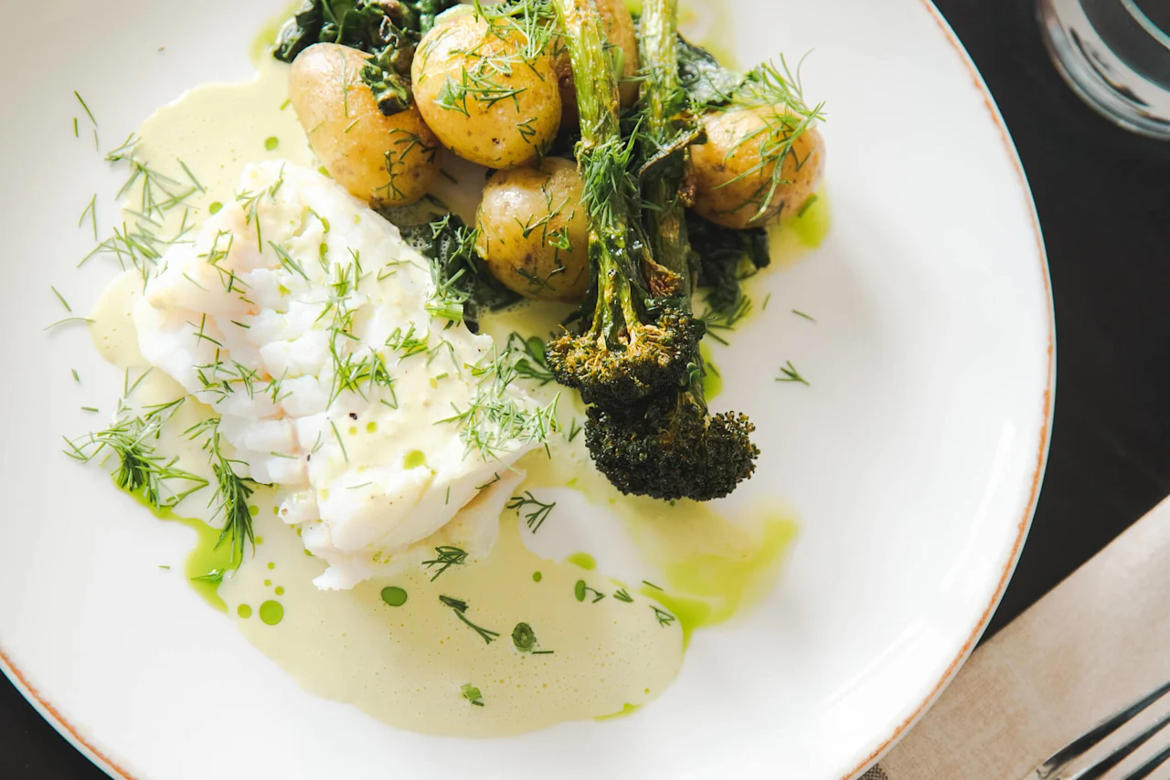 Panert hvit fisk med poteter og brokkoli toppet med fersk dill på en hvit tallerken, omgitt av en lysegrønn saus, pyntet med grønne urter.