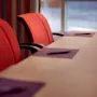 Punaisia toimistotuoleja neuvottelupöydän ympärillä muistilehtiöineen, sisätilassa suurten ikkunoiden lähellä.