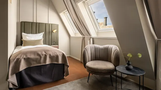 Et lille, moderne soveværelse har en seng med en grøn sengegavl og brunt sengetøj. Der er en blød stol og et lille bord med en vase ved siden af skråvinduer.