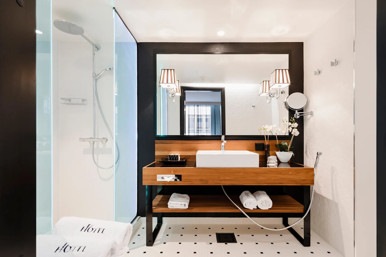 Modern bathroom with a wood-framed sink and mirror, adjacent to a glass shower. White towels are labeled "HOTEL". The setting features elegant lighting and orchid decor.