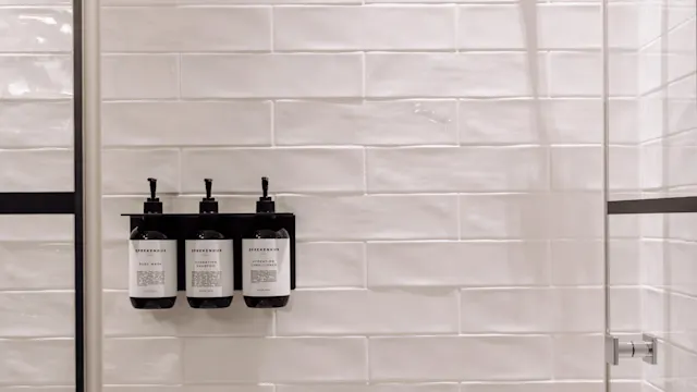 Tre flasker med pumper er montert på en hvit flislagt dusjvegg. De er merket "SPREKENHUS" med produkter: "BODY WASH," "HYDRATING SHAMPOO," og "HYDRATING CONDITIONER."