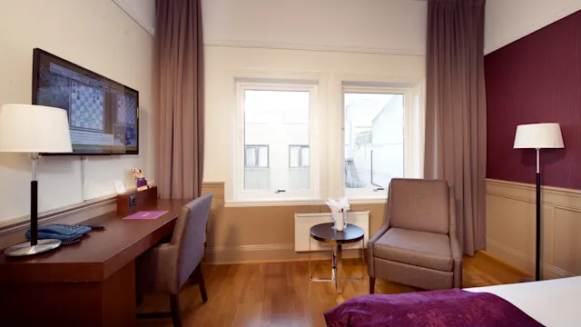A hotel room interior with a large bed, a desk with a television, a chair, and windows looking onto a building.