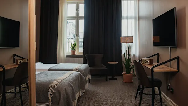 To pent oppreide enkeltsenger med gråe pledd er plassert i et hotellrom med en veggmontert TV, mørke gardiner, et lite skrivebord, stol og store potteplanter.