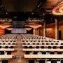 Rows of tables and chairs are arranged in a large, modern conference hall with wooden walls, red lighting accents, and circular chandeliers. A large, empty screen is displayed at the front.