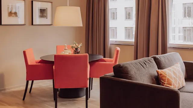Round table with red chairs holds flowers and a bottle, in a warmly lit room with framed art on beige walls and a gray sofa by windows.