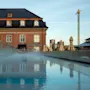 Et tåget basseng reflekterer en historisk murbygning med arkade vinduer, omgitt av lukkede gulstripete parasoller i et urbant miljø med et høyt, dekorativt tårn i bakgrunnen.