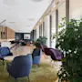 Komfortable loungestoler omgir et lite bord på et grønt teppe; rommet åpner seg inn i et moderne, romslig rom med høye vinduer, mange planter og en lang bardisk med krakker.