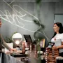 A man with a suitcase speaks to a smiling receptionist in a modern hotel lobby, featuring elegant decor and lighting. The receptionist is wearing a t-shirt with text partially visible: “strawberry.”