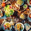 Bowls and plates filled with various salads, soups, and breads are arranged on a dark marble table, surrounded by cutlery, napkins, and glasses, creating a colorful dining setting.