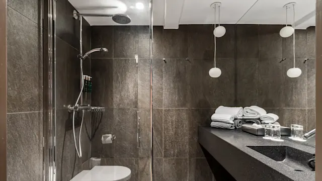 A glass-enclosed shower with chrome fixtures stands beside a dark stone wall. Nearby, folded towels rest on a countertop under soft lighting in a modern bathroom setting.