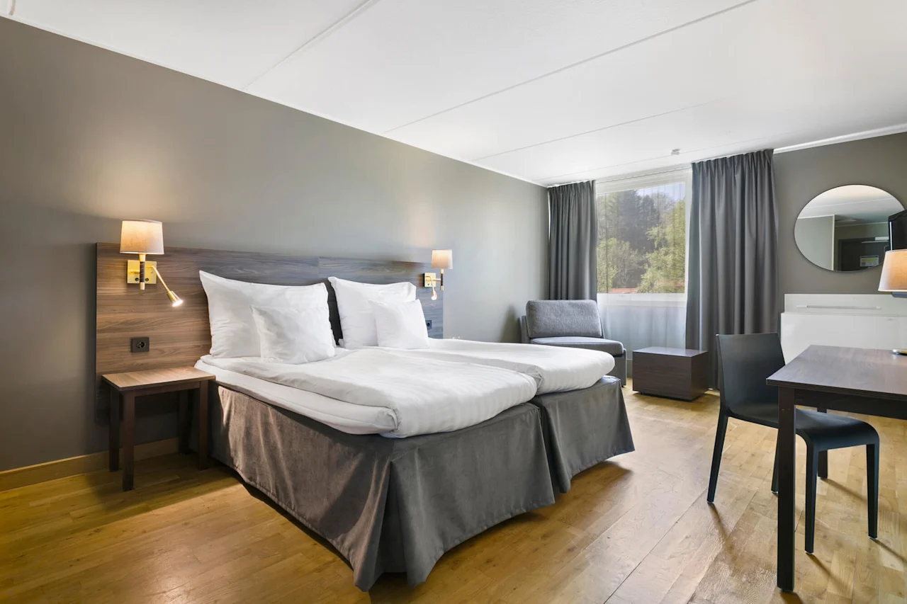 En säng med vita lakan står bredvid ett litet bord med lampa i ett modernt hotellrum. Stort fönster med gardiner och skogsutsikt, stol och skrivbord i bakgrunden.