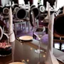 A restaurant table is set with wine glasses, folded napkins, cutlery, and a floral centerpiece. The background features multiple elegant dining chairs under soft lighting.